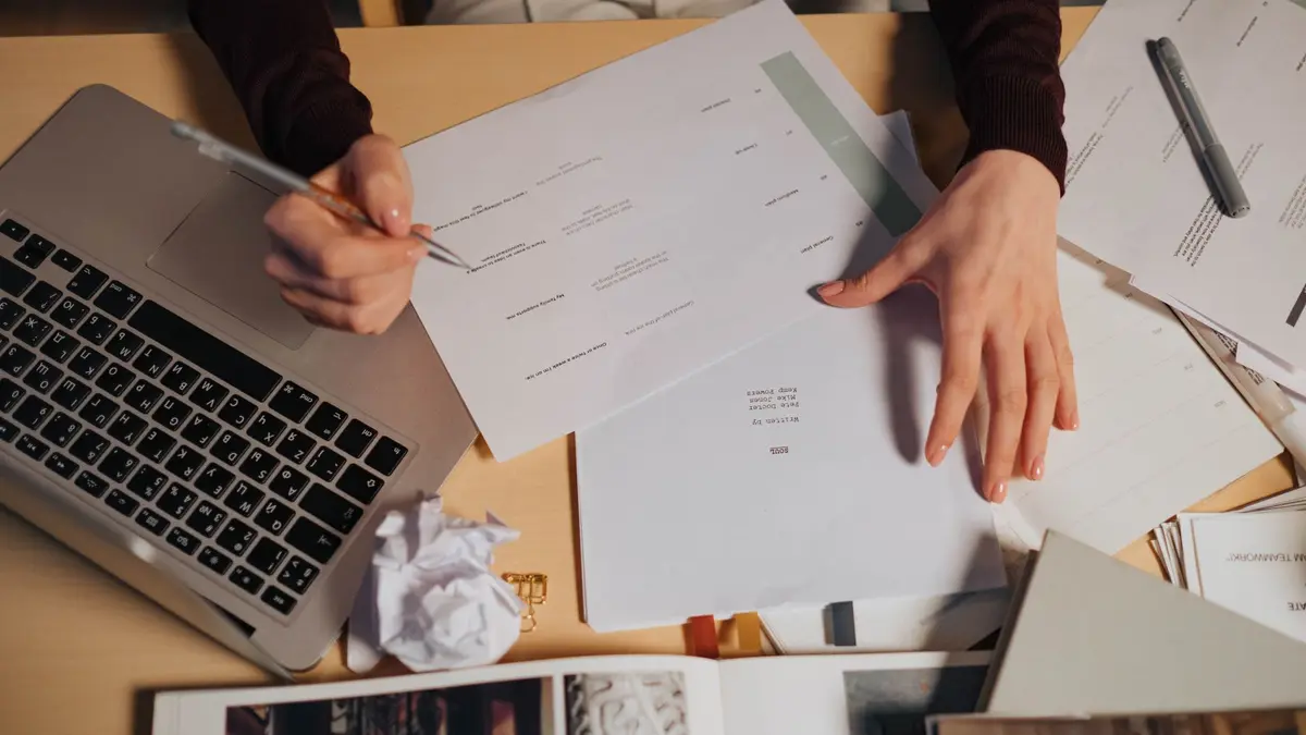 Pessoa revisando anotações e documentos ao lado de um notebook